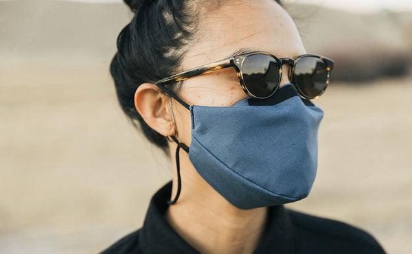 Person standing outdoors in a grassy field wearing the V6 Mask in Pacific Blue. The person is wearing sunglasses.